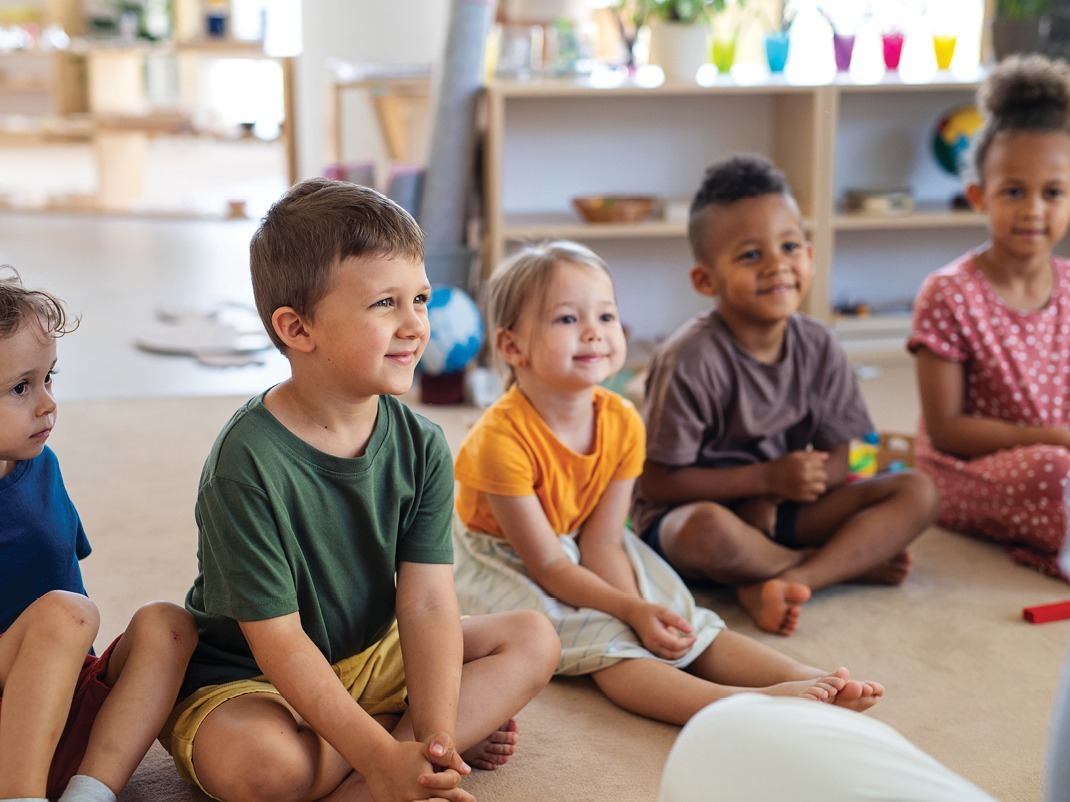 five young children sitting on a soft carpet and smiling at their instructor